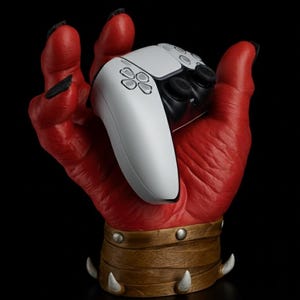 Peut inclure: Un support de manette de jeu en forme de main rouge texturée, avec un bracelet marron et doré et des griffes blanches. Le support tient une manette de jeu blanche et noire, sur fond noir.