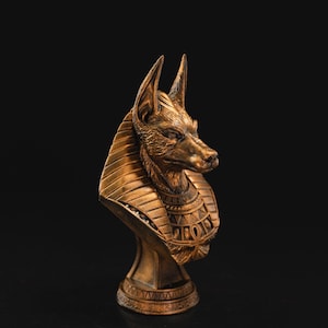 Könnte beinhalten: Eine bronzefarbene Büste von Anubis, dem altägyptischen Gott des Jenseits, mit dem Kopf eines Schakals. Die Skulptur weist detaillierte Schnitzereien des Fells, der Ohren und der Gesichtszüge des Schakals sowie den traditionellen Kragen und Kopfschmuck auf.