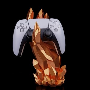 Puede incluir: Un mando de videojuegos blanco y negro descansa sobre un soporte dorado en forma de mano geométrica. El soporte tiene un diseño similar a un cristal. El fondo es negro, resaltando el mando y el soporte.