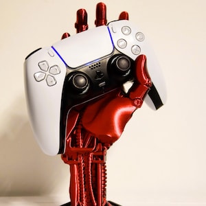 Könnte beinhalten: Ein weiß-schwarzer Videospiel-Controller wird von einer roten Roboterhand gehalten. Der Controller hat ein blaues Licht und verschiedene Tasten. Die Hand ist auf einem schwarzen sechseckigen Sockel montiert.