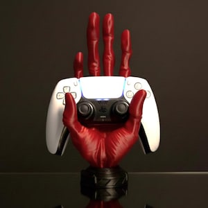 Könnte beinhalten: Ein roter, handförmiger Controller-Ständer, der einen weißen und schwarzen Gamecontroller hält. Der Ständer hat einen schwarzen Sockel und ist so konzipiert, dass er den Controller aufrecht hält. Der Controller hat ein blaues Licht.
