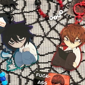 May include: A collection of anime-themed merchandise including two acrylic keychains featuring characters with dark hair, a beaded bracelet with the name KIRA, a blue and black beaded bracelet with a heart, a rosary, and a rectangular card with text.
