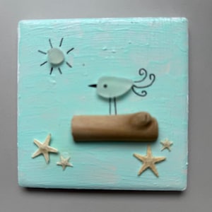 Peut inclure: Plaque carrée en bois peint en turquoise, représentant une scène de plage fantaisiste. Un oiseau en verre perché sur une bûche en bois, avec des accents de soleil et d'étoiles de mer. L'œuvre évoque un thème côtier.