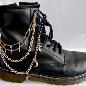Puede incluir: Una bota de cuero negro con cordones que presenta un arnés de cadenas decorativo en tono oro rosa. El accesorio incluye varias cadenas drapeadas, dijes de barras verticales delgadas y pequeños colgantes de media luna en el lateral.