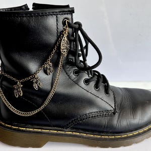 Pode incluir: Uma bota de cano curto preta com atacadores frontais e fecho lateral. A bota é adornada com um acessório de corrente de metal dourado com pequenos pingentes em forma de folha e coruja presos aos ilhós e à aba traseira.