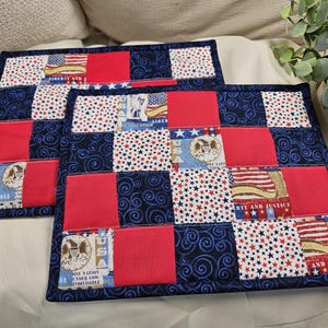 May include: Two rectangular placemats with a patchwork design. The design incorporates red squares, navy blue fabric with white swirls, and fabric with American flag and star patterns. The edges are finished with a navy blue border.