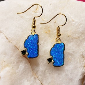 May include: A pair of gold earrings featuring a blue opal shaped like the state of Michigan with a small black gemstone accent.