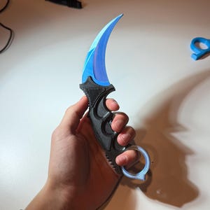 May include: A black and blue karambit knife with a curved blade. The blade has a gradient of blue hues. The handle is black with a finger ring. The knife is held in a hand.