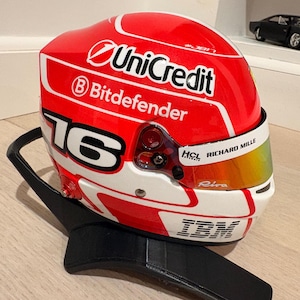 May include: A red and white racing helmet with sponsor logos, including "UniCredit" and "Bitdefender." The helmet features the number "16" and a gold visor. It rests on a black stand.