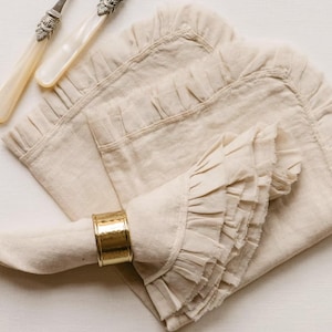 May include: Beige linen napkins with ruffled edges and a gold napkin ring. Two antique-style knives with ornate handles are also visible. The napkins are folded and arranged on a light-colored surface, creating a soft, elegant aesthetic.