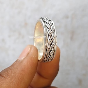 Puede incluir: Un anillo de plata con un diseño trenzado. El anillo tiene un patrón tejido texturizado alrededor de la banda. El anillo se sostiene entre dos dedos sobre un fondo borroso. El texto "MKJEWELLERSGIFT" es visible en el anillo.