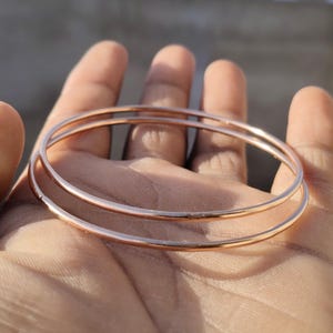 Könnte beinhalten: Drei dünne Armreifen aus Roségold werden gezeigt. Die Armreifen sind kreisförmig und haben eine glatte, polierte Oberfläche. Der Schmuck wird in einer Hand gehalten, wobei der Hintergrund unscharf ist.