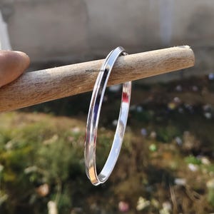 May include: A polished silver bangle bracelet. The smooth surface reflects light, giving a bright appearance. The round bracelet is made of metal. The bangle is resting on a wooden dowel. The background is blurred.