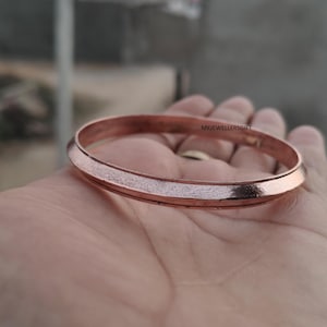 May include: A copper-colored bangle bracelet with a textured center band. The bracelet is round and smooth on the edges. The text "MKJEWELLERSGIFT" is visible on the bracelet. The bracelet is held in an open hand.