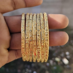 Puede incluir: Un conjunto de siete pulseras doradas con intrincados diseños florales y de hojas. Las pulseras están apiladas, mostrando su diseño detallado y acabado brillante. Las joyas se presentan sobre un fondo borroso.