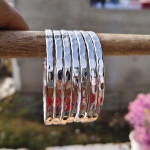 May include: A set of seven silver bangles with a hammered texture. The bangles are displayed on a wooden dowel, with the text "MKJEWELLERSGIFT" visible. The background is blurred, with hints of green and pink.