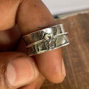 Peut inclure: Une bague spinner argentée avec une surface texturée. La bague présente un motif en forme de cœur et d'autres détails en relief. La bague est tenue entre les doigts.