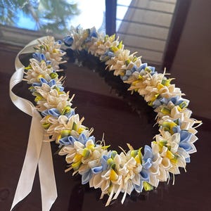 May include: A lei, a traditional Hawaiian garland, made of white, blue, and yellow fabric flowers. The lei is tied with a white ribbon. The flowers are arranged in a circular pattern.