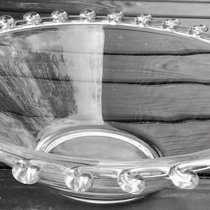 May include: Clear glass serving bowl with a decorative rim of small, round glass accents. The bowl has a wide, shallow design and a central base. The bowl is sitting on a wooden surface.