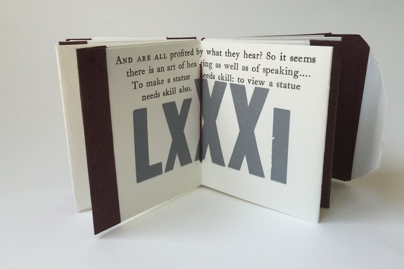 May include: A small, white book with a brown spine. The book is open to reveal a page with the text "AND ARE ALL profited by what they hear? So it seems there is an art of hearing as well as of speaking.... To make a statue needs skill also. needs skill: to view a statue LXXXI" printed in black. The page is decorated with a large, gray number "LXXXI" printed in a bold font.