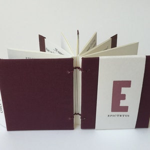 May include: A small, burgundy-colored book with the text "Once Said a Stoic" on the front cover. The book is open to reveal a white page with a large, pink letter "E" and the word "EPICTETUS" printed below it.