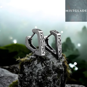 Può includere: Un paio di orecchini a cerchio argentati con un design geometrico. Ogni orecchino presenta una forma rettangolare con un motivo decorativo in bianco e nero. Gli orecchini sono esposti su una roccia muschiosa.