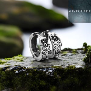 May include: A pair of silver-colored hoop earrings with a wolf head design. The earrings are detailed with black patterns and sit on a mossy rock. The word "MISTGLADE" is visible in the background.