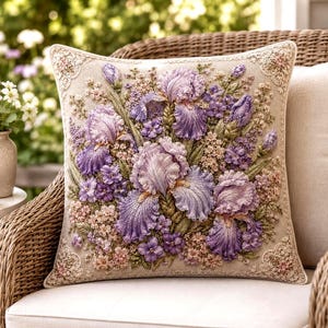 May include: A square throw pillow featuring a beige fabric base with intricate, raised embroidery of purple and lavender iris flowers, buds, and small accent blossoms. The corners are decorated with delicate, cream-colored lace trim.