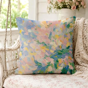 May include: Square decorative pillow with an abstract floral design. The pillow features a blend of pastel colors, including pink, blue, yellow, and green, creating a soft, artistic aesthetic. The pillow is placed on a floral patterned cushion.