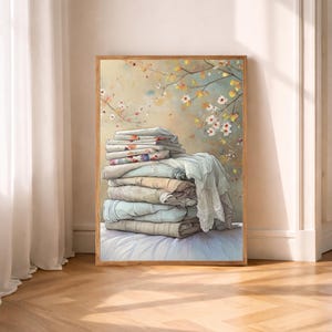 May include: A framed art print depicts a stack of folded linens in soft pastel colors. The linens are arranged on a white surface, with a backdrop of a floral scene in muted tones. The artwork is in a wooden frame.