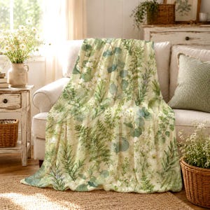 May include: A soft throw blanket featuring a botanical print with various green leaves, ferns, and small white flowers on a light cream background. The pattern covers the entire surface of the fabric, displaying a detailed arrangement of foliage.