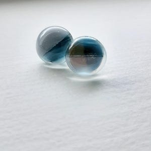 Peut inclure: Deux billes de verre transparentes avec des motifs internes bleus et bruns. Les billes sont sphériques et reposent sur une surface blanche. La lumière se reflète sur le verre, créant des reflets et des ombres.