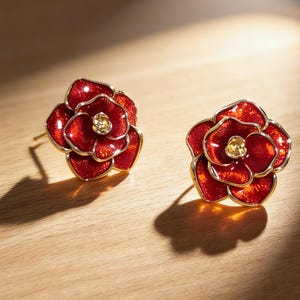 May include: A pair of red flower stud earrings with gold-coloured edging and a yellow centre stone. The earrings are displayed on a light wooden surface, with shadows from a light source.