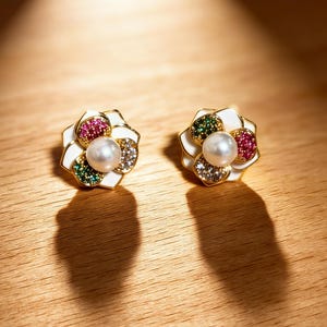 May include: A pair of gold-toned flower-shaped earrings. Each earring features a white enamel base, a central pearl, and colorful gemstones in red, green, and clear. The earrings are set on a wooden surface.