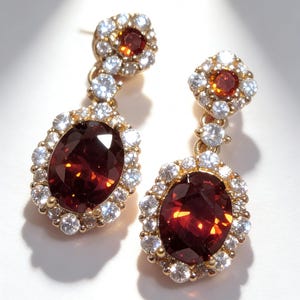 May include: A pair of gold-coloured drop earrings. Each earring features a large, oval, deep red gemstone surrounded by a halo of small, clear gemstones. The top of each earring has a smaller, square-shaped cluster of gemstones.