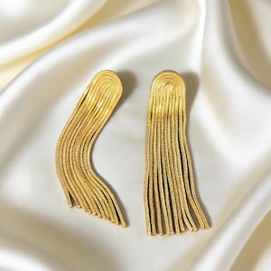 May include: A pair of gold tassel earrings. Each earring has a curved top with vertical, dangling strands. The earrings are presented on a white, silky background.