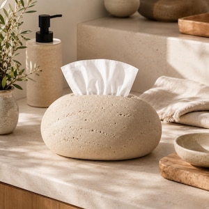 Puede incluir: Una funda para caja de pañuelos de color beige y forma ovalada con una superficie texturizada y porosa que recuerda a la piedra natural. La funda presenta una abertura horizontal central por la que se dispensan pañuelos blancos.