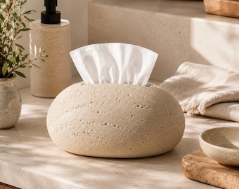 May include: A beige, oval-shaped tissue box cover with a textured, porous surface resembling natural stone. The cover features a central horizontal opening from which white tissues are dispensed, providing a functional and neutral home accessory.
