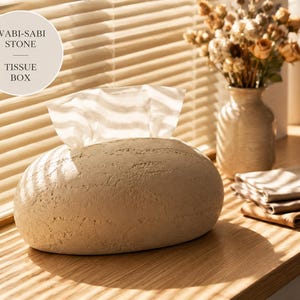 May include: A beige, oval-shaped tissue box with a textured, stone-like surface. The container features a central opening at the top from which white tissues are dispensed. The text WABI-SABI STONE TISSUE BOX is visible in a circular graphic.