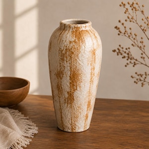 May include: A tall ceramic vase with a textured surface featuring a mottled pattern of cream and warm ochre brown. The vessel has a narrow neck, a rounded body that tapers towards the base, and a distinctively rough, uneven finish throughout.