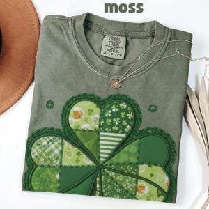 May include: A moss green t-shirt with a large, appliqued shamrock design in various shades of green. The shamrock features a detailed, patchwork design with beer mugs and clover patterns. A silver necklace with a small pendant rests on the shirt.