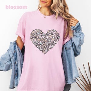 May include: A light pink short-sleeve t-shirt featuring a large heart-shaped graphic composed of various small illustrations of coffee pots, cups, and croissants in shades of brown, blue, and white. The word blossom is printed in the top left corner.