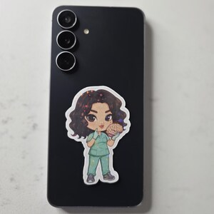 Puede incluir: Una pegatina de una enfermera de dibujos animados sosteniendo un cerebro, colocada en un tel&eacute;fono inteligente negro. La enfermera lleva un uniforme verde y tiene el pelo rizado oscuro. La pegatina tiene un borde blanco y un fondo gris claro.