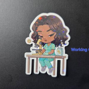 Puede incluir: Una pegatina hologr&aacute;fica con una ilustraci&oacute;n de dibujos animados de una persona con uniforme turquesa usando un microscopio. El texto "Working Girl 1" es visible.