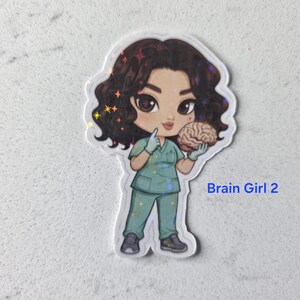 Puede incluir: Una pegatina con una ilustraci&oacute;n de dibujos animados de una mujer con uniforme turquesa sosteniendo un cerebro. La mujer tiene el pelo rizado oscuro y lleva guantes. El texto "Brain Girl 2" es azul.
