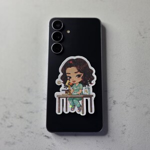 Puede incluir: Un tel&eacute;fono inteligente negro con una pegatina de una cient&iacute;fica de dibujos animados examinando un microscopio. La pegatina muestra a una mujer con cabello casta&ntilde;o con una bata y pantalones verdes. El tel&eacute;fono tiene tres lentes.