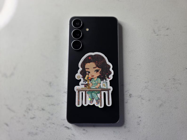 Puede incluir: Un tel&eacute;fono inteligente negro con una pegatina de una cient&iacute;fica de dibujos animados examinando un microscopio. La pegatina muestra a una mujer con cabello casta&ntilde;o con una bata y pantalones verdes. El tel&eacute;fono tiene tres lentes.