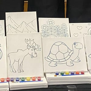 May include: A collection of small, white canvas boards with line drawings of a rocket, mermaid, and shark. Each canvas has a small paint palette. The canvases are ideal for painting and creative activities.