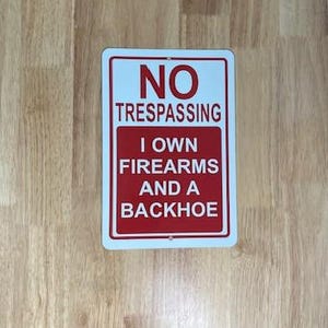 May include: Rectangular sign with a white background and red border. The sign reads "NO TRESPASSING" at the top, and below that, "I OWN FIREARMS AND A BACKHOE" in red text.