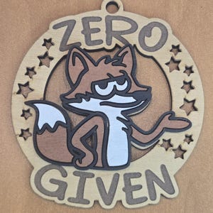 May include: A circular wooden ornament featuring a cartoon fox with a skeptical expression. The text ZERO GIVEN is engraved above and below the fox. The design includes small star cutouts along the sides and a hole at the top for hanging.